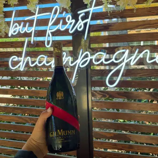 A bottle of GH Mumm held up against a sign that says but first champagne