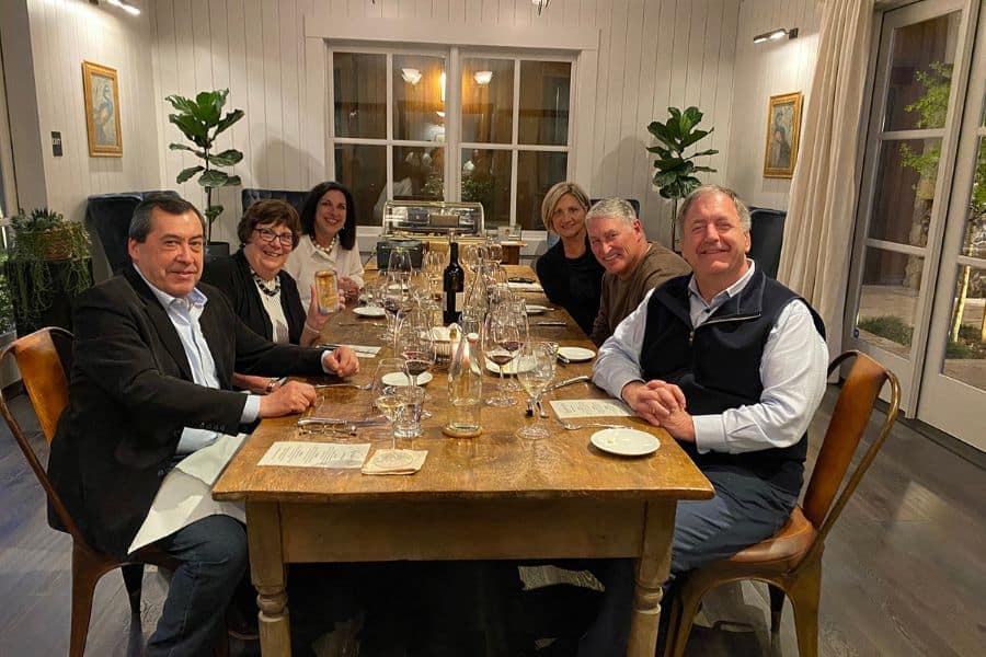 Six people around a large wood table with wine smiling at the camera