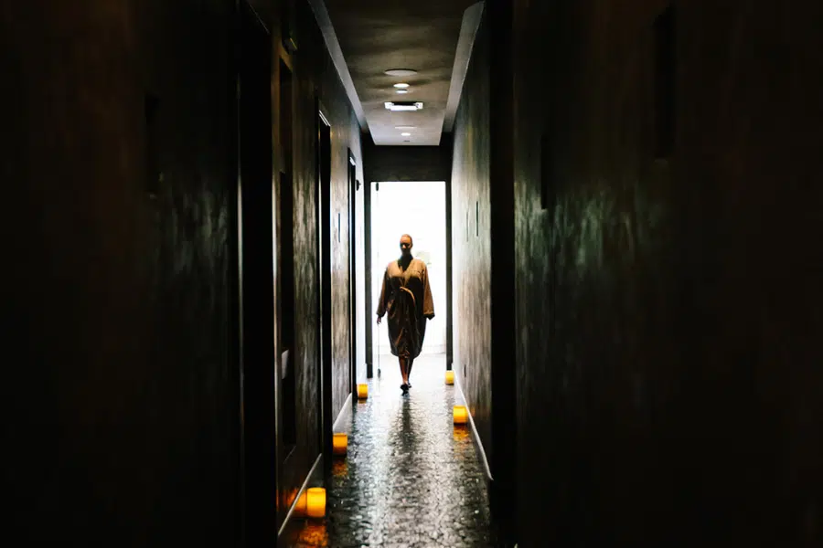 A dimly lit spa hallway with candles on the ground with a woman walking down the hallway in a spa robe