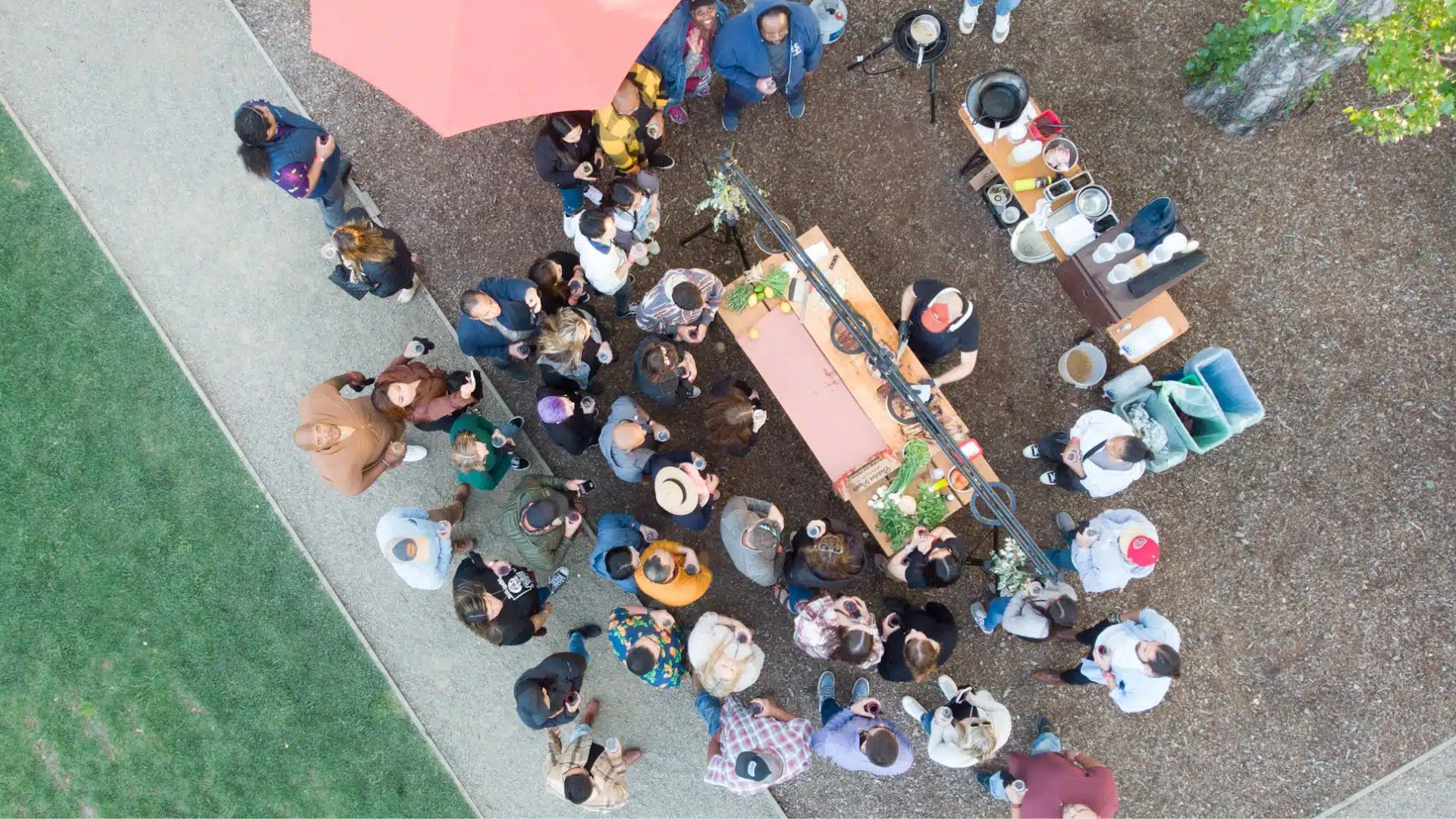 Aerial view of outdoor Yountville wine event with people gathered around tasting table under umbrella and string lights.